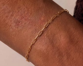 May include: A delicate gold chain bracelet featuring elongated, rectangular links connected in a continuous, repeating pattern. The metal has a polished, reflective finish that catches the light, creating a simple and refined appearance on the wrist.