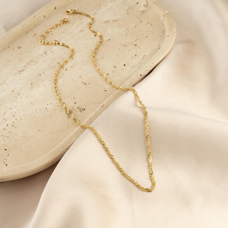 Op de afbeelding: Een gouden ketting met een delicaat, gedraaid touwontwerp. De ketting wordt getoond op een lichtbeige oppervlak.