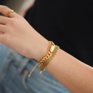 May include: A gold chain bracelet with a thick, chunky link chain and a smaller, delicate chain. The bracelet is layered with a third chain that has a rectangular, flat piece in the center.