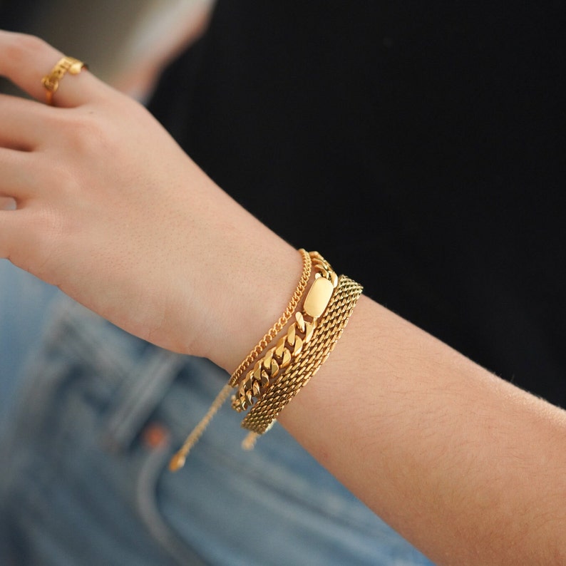 May include: A gold chain bracelet with a thick, chunky link chain and a smaller, delicate chain. The bracelet is layered with a third chain that has a rectangular, flat piece in the center.