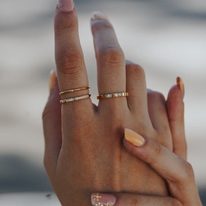 Puede incluir: Una mano con tres anillos de oro con peque&ntilde;os diamantes en el dedo medio. Las u&ntilde;as est&aacute;n pintadas con un esmalte naranja claro y una u&ntilde;a tiene un dise&ntilde;o de flor blanca y amarilla.
