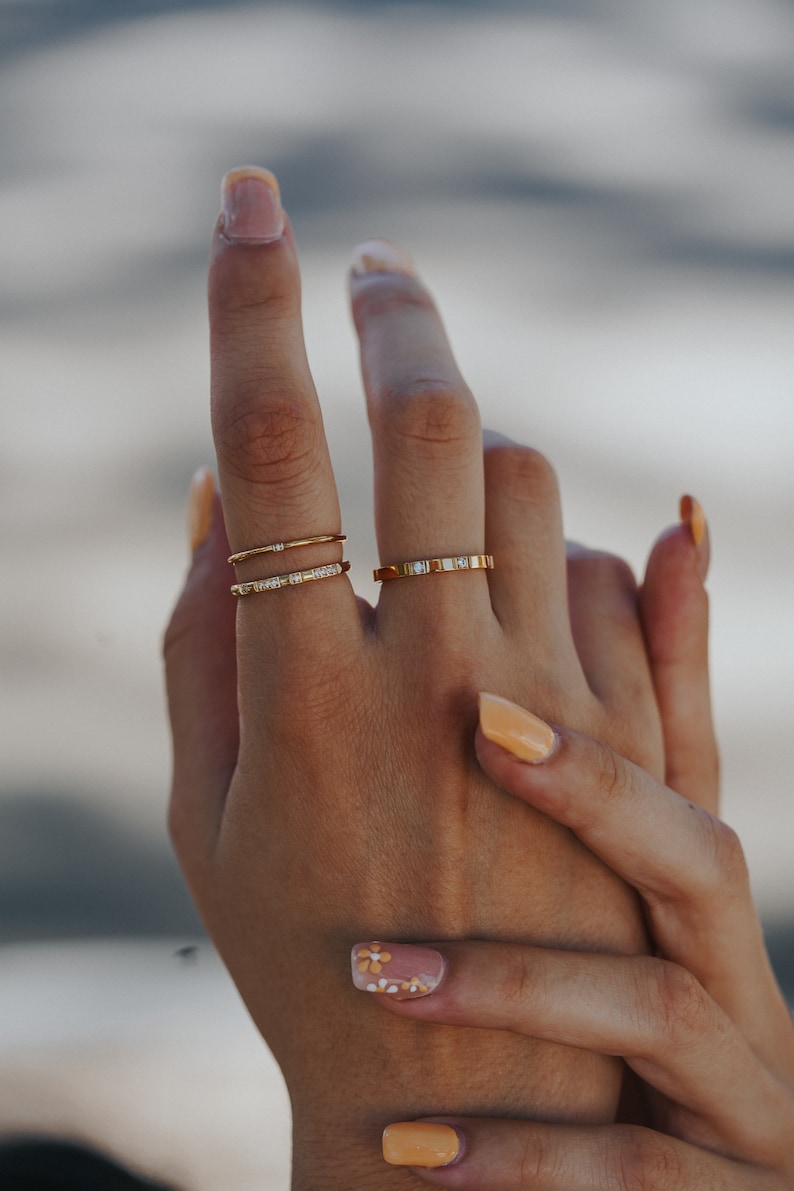 Puede incluir: Una mano con tres anillos de oro con peque&ntilde;os diamantes en el dedo medio. Las u&ntilde;as est&aacute;n pintadas con un esmalte naranja claro y una u&ntilde;a tiene un dise&ntilde;o de flor blanca y amarilla.