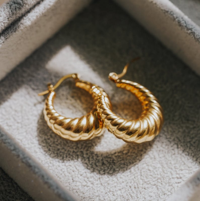 May include: A pair of gold-coloured hoop earrings with a textured, ridged design. The earrings are displayed inside a grey jewellery box, catching the light and showcasing their shiny finish. The hoops have a classic, elegant style.