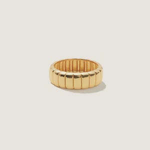 May include: A gold-colored ring with a ribbed design. The ring has a thick band with vertical ridges around the entire circumference. The ring is made of a shiny, polished metal and sits on a white background.