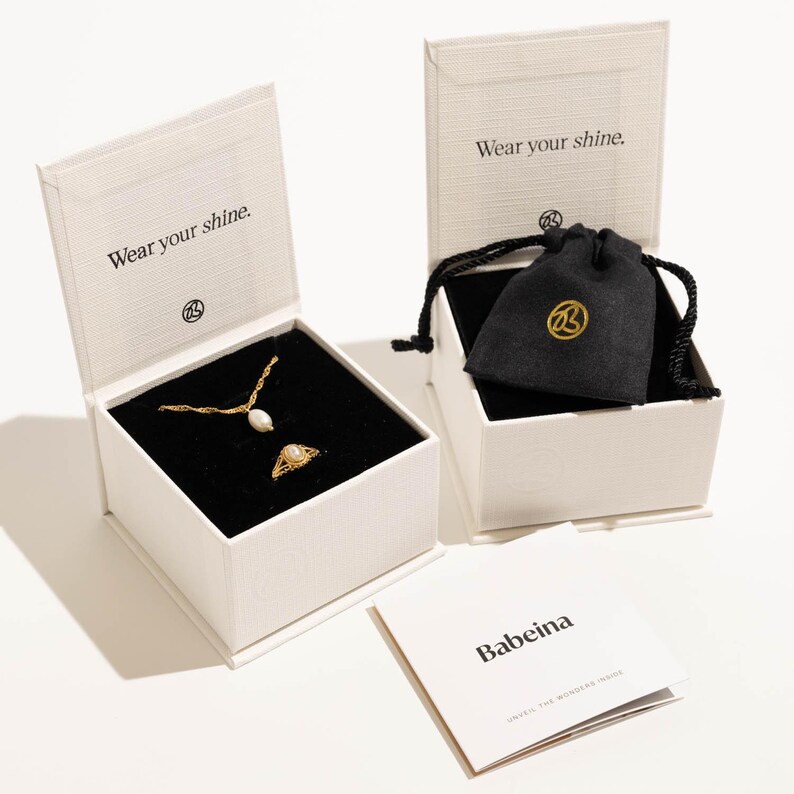 May include: A white box with the text "Wear your shine." on the lid. Inside the box is a gold necklace with a pearl pendant and a gold ring. The box is sitting on a white surface. There is a smaller white box with a black velvet bag inside. The bag has a gold logo on it. The text "Wear your shine." is on the lid of the smaller box. There is a white card with the text "Babeina" on it.