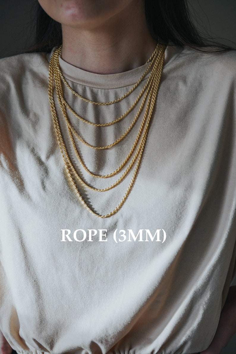 May include: A close-up of a person wearing a beige t-shirt with a layered gold rope chain necklace. The necklace is 3mm thick and has a shiny finish. The text "ROPE (3MM)" is printed on the shirt.