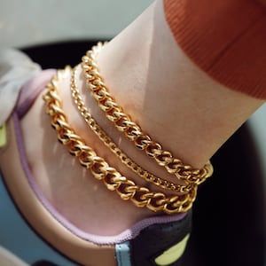 Peut inclure: Trois bracelets de cheville dorés de différentes épaisseurs ornent une cheville. Les chaînes sont superposées et portées sur une basket aux couleurs beige, bleu et noir. Les bijoux ont une finition polie et réfléchissent la lumière.