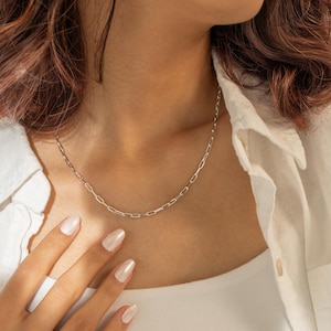 Op de afbeelding: Een zilveren ketting met een delicaat paperclip-kettingontwerp. De ketting wordt gedragen door een persoon met een wit shirt.