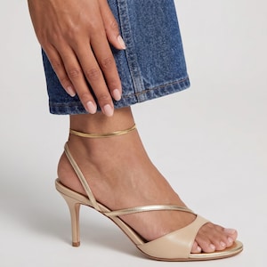 May include: A gold ankle bracelet and beige leather sandals with thin straps and a stiletto heel. The sandals have a toe strap and a strap that crosses the foot. The ankle bracelet is a simple, flat design.