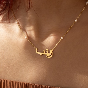 May include: A gold necklace with a delicate chain and small pearl accents. The necklace features a personalized nameplate with the Arabic word "Layla" in a stylish script.