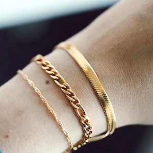 May include: A close-up of a wrist adorned with three gold bracelets. The bracelets include a delicate twisted chain, a curb chain, and a flat, herringbone-style bracelet. The jewelry is displayed against a skin tone background.