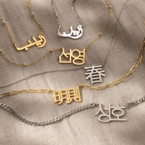 May include: A collection of gold and silver necklaces with personalized nameplates featuring Arabic, Korean, and Chinese characters. The necklaces are displayed on a textured fabric background.