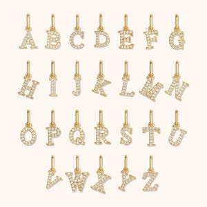 May include: A collection of gold-colored alphabet charms, each letter encrusted with small, sparkling stones. The charms are suspended from small loops, creating a complete alphabet set. The background is a soft, neutral color.