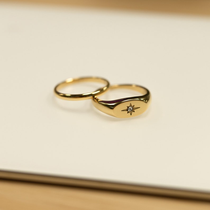 May include: Two gold rings on a white surface. One is a simple band, and the other is a signet ring with a starburst design and a small, clear stone. The rings are reflective and appear to be made of polished metal.