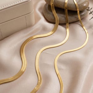 May include: A gold-toned herringbone chain necklace featuring a flat, flexible, and tightly woven link design. The metallic surface reflects light, showcasing a smooth, continuous pattern that curves gracefully across the frame.