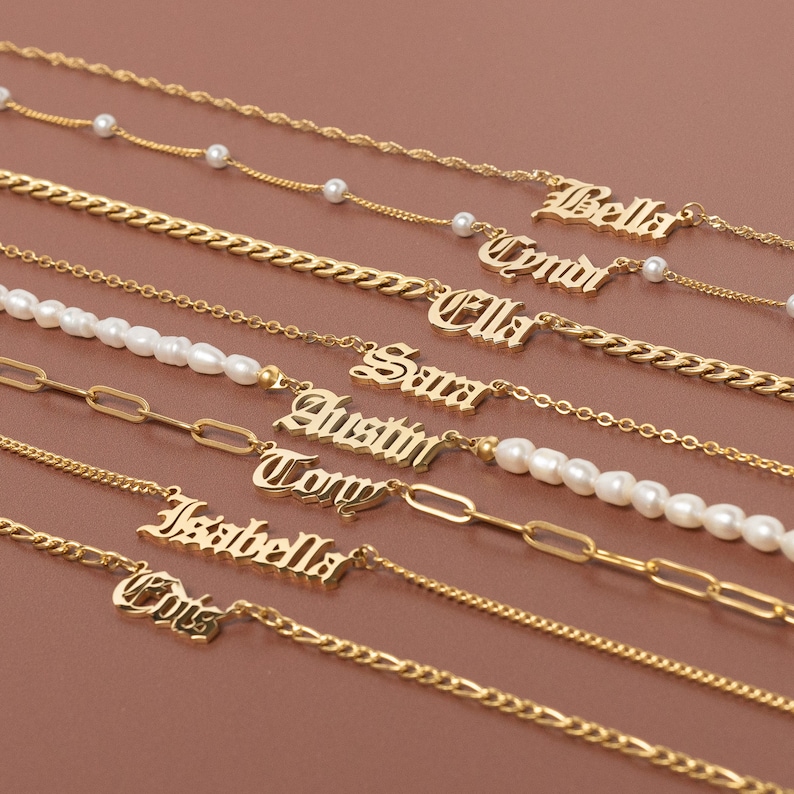 K&ouml;nnte beinhalten: Ein Set aus neun goldenen Halsketten mit verschiedenen Namen in gotischer Schrift. Jede Halskette hat einen anderen Kettenstil, darunter eine zarte Kette, eine Curb-Kette und eine Kabelkette. Einige Halsketten haben Perlenakzente.