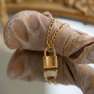 May include: A gold chain necklace with a small gold padlock pendant. The necklace is lying on a piece of driftwood.