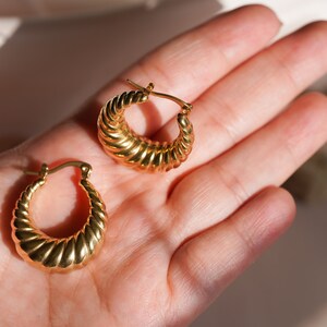 May include: A pair of gold-coloured hoop earrings. The earrings have a ridged, croissant-like design. The earrings are displayed on a light-toned background.