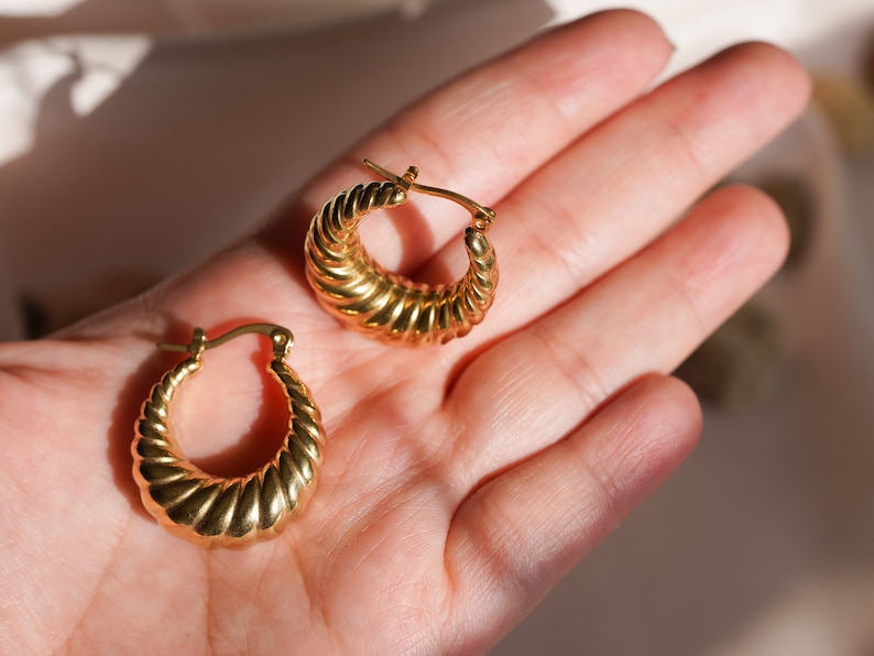May include: A pair of gold-coloured hoop earrings. The earrings have a ridged, croissant-like design. The earrings are displayed on a light-toned background.