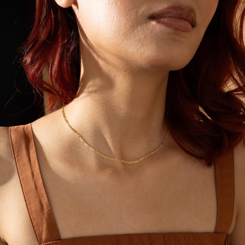 K&ouml;nnte beinhalten: Eine goldene Kettenkette mit einem zarten, leicht geschwungenen Design. Die Kette wird von einer Person mit roten Haaren getragen, aber der Fokus liegt auf der Kette selbst.