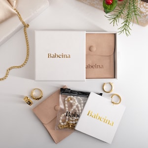 May include: A white box with the text "Babeina" on it. The box is open and contains a small brown pouch with the text "Babeina" on it. There are also two gold hoop earrings, a gold chain necklace, and a pearl necklace in a clear plastic bag.