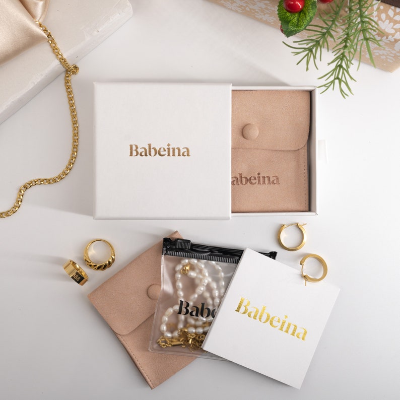 May include: A white box with the text "Babeina" on it. The box is open and contains a small brown pouch with the text "Babeina" on it. There are also two gold hoop earrings, a gold chain necklace, and a pearl necklace in a clear plastic bag.