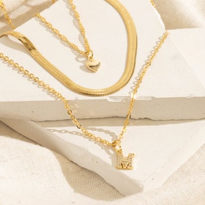 May include: Three gold necklaces. The top necklace is a herringbone chain. The middle necklace has a heart pendant. The bottom necklace has a pendant with the letter "M" encrusted with small, clear stones. The necklaces are displayed on a white surface.