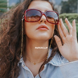 May include: Large, square-framed sunglasses with a dark red hue and a gold chain. The chain is attached to the glasses and hangs down. The word "PAPERCLIP" is printed on the image.