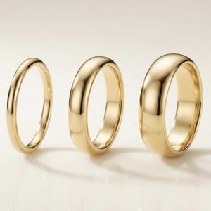 May include: Three gold wedding bands of varying widths are displayed. The rings have a polished, reflective surface, with the light creating highlights and shadows. The bands are set against a soft, neutral background, emphasizing their simple elegance.