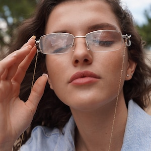 May include: Silver-framed eyeglasses with a gold chain. The glasses have a rectangular shape and are being held up by a person. The chain is thin and delicate, adding a touch of elegance to the eyewear.