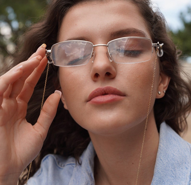 May include: Silver-framed eyeglasses with a gold chain. The glasses have a rectangular shape and are being held up by a person. The chain is thin and delicate, adding a touch of elegance to the eyewear.