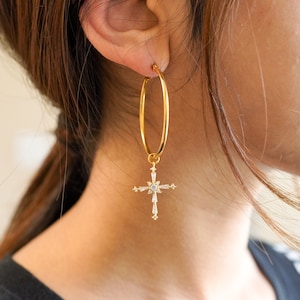 May include: Gold-tone hoop earrings with a dangling cross pendant. The cross is adorned with clear, sparkling crystals. The earrings are a classic and elegant accessory.