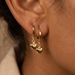 May include: Close-up of gold hoop earrings with heart-shaped charms. The earrings are hanging from a person's ear. The heart charms are smooth and shiny, reflecting light. The earrings are a simple and elegant design.