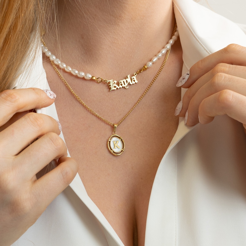 K&ouml;nnte beinhalten: Eine goldene Kettenkette mit einer dar&uuml;berliegenden Perlenkette. Die goldene Kette hat einen Anh&auml;nger mit einem wei&szlig;en Kreis und dem Buchstaben "K" in Gold. Die goldene Kette hat auch ein Namensschild mit dem Namen "Kayla" in Gold.
