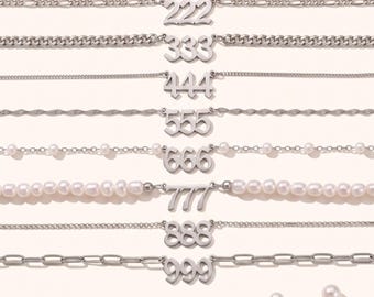 May include: A collection of eight silver-toned necklaces arranged horizontally, each featuring a unique three-digit number pendant from 222 to 999. The chains vary in style, including link, cable, and pearl-accented designs, with loose pearls nearby.