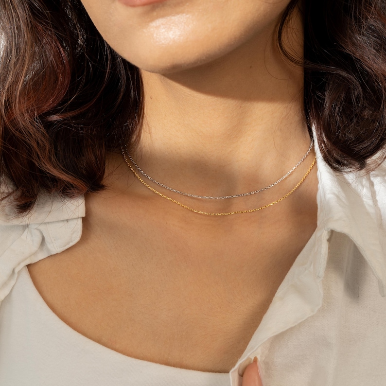 Puede incluir: Primer plano de dos delicados collares. Uno es plateado y el otro dorado. Los collares est&aacute;n superpuestos y descansan sobre el cuello de una persona, contra una camisa blanca. La iluminaci&oacute;n es suave, resaltando los sutiles detalles de la joyer&iacute;a.