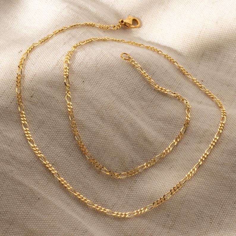 K&ouml;nnte beinhalten: Zwei goldene Kettenhalsb&auml;nder, eines l&auml;nger als das andere, liegen auf einer beigefarbenen Stoffoberfl&auml;che.