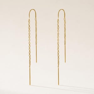 May include: A pair of gold threader earrings featuring a delicate chain design. Each earring consists of a thin, curved wire that transitions into a fine link chain, ending in a straight, smooth metal post for a sleek and elongated appearance.
