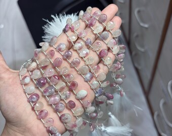 May include: Strands of pink and clear teardrop-shaped gemstones, strung together with silver wire. The gemstones are held in a hand, with white tassels at the ends of the strands. The gemstones have a translucent quality.