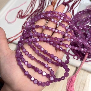 May include: Several strands of faceted, deep purple gemstone beads. The beads are hexagonal in shape and strung together. A pink tassel and red string are visible in the background. The beads are held in a hand.