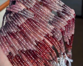 May include: Strands of small, faceted gemstones in shades of pink, red, and clear. The beads are strung together, with white tassels at the ends. The gemstones are held in a hand, showcasing their color variations and sparkle.