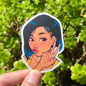 May include: A sticker featuring a cartoon illustration of a woman with blue hair, wearing a yellow shirt and giving a peace sign. She has stars in her hair and a ring on her finger.