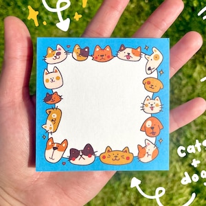 May include: A square blue notepad with a border of cartoon cats and dogs. The notepad has the words "Note Pad!" written at the top and "cats + dogs" at the bottom. The notepad is held in a hand against a green background.