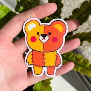 May include: A sticker of a cartoon teddy bear with a patchwork design. The bear is orange and yellow with red stitching. The bear is smiling and has a cute expression.