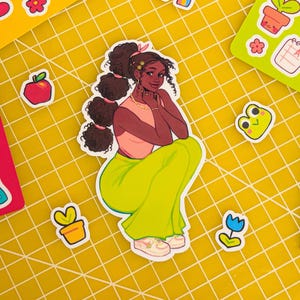 May include: A sticker of a cartoon woman with brown hair, wearing a pink shirt and green pants. She is sitting with her legs crossed and has a flower in her hair.