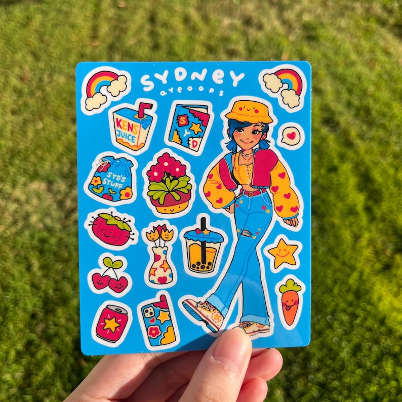May include: A sticker sheet with a blue background featuring a cartoon character wearing a yellow hat, blue jeans, and a red and yellow jacket. The sticker sheet also includes a variety of other stickers, including a rainbow, a can of soda, a flower, and a cup of boba tea. The text "SYDNEY aveooops" is at the top of the sheet.