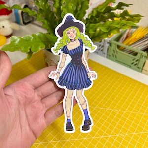 May include: A sticker of a cartoon witch with green hair, wearing a black and blue dress, a witch's hat, and black shoes. The witch has a stitched-up appearance and is smiling.