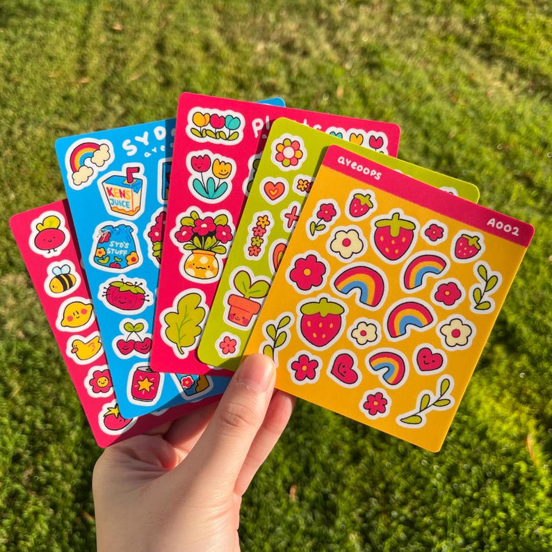 May include: Four sheets of colorful stickers with various designs, including strawberries, rainbows, flowers, and plants. The stickers are on a yellow, pink, and blue background. The text on the stickers reads "ayeoops" and "A002".