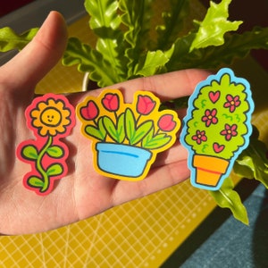 Puede incluir: Tres pegatinas coloridas con plantas en maceta. La primera pegatina muestra una flor amarilla con una cara sonriente en un tallo rojo. La segunda pegatina muestra una planta en maceta con flores rojas y hojas verdes. La tercera pegatina muestra una planta en maceta con hojas verdes y corazones rojos.