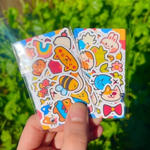 May include: A clear plastic bag filled with colourful stickers featuring cartoon animals, flowers, and geometric shapes. The stickers are on a white background.